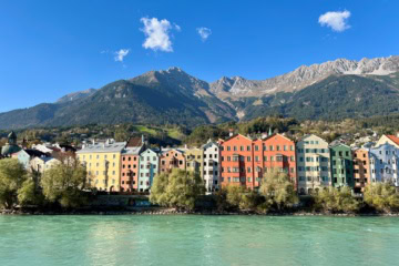 Blick über den türkisfarbenen Inn auf die Häuserzeile von Mariahilf in Innsbruck mit Fassaden in Gelb, Rot, Grün und Blau. Dahinter steigt der bewaldete Hang zur Nordkette auf, über den Felswänden steht blauer Himmel.