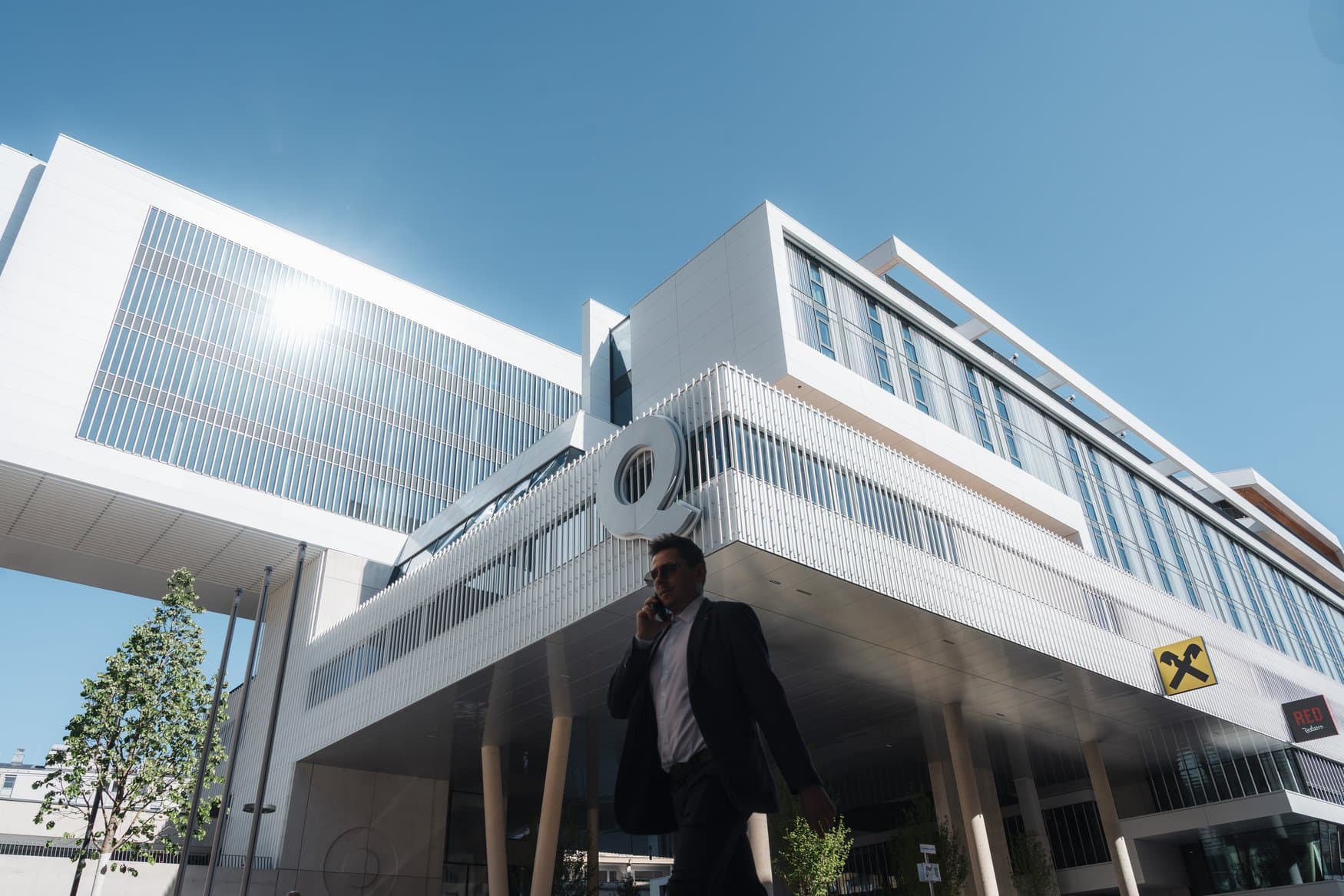 Untersicht auf das Gebäude Raiqua in Innsbruck mit weißer Lamellenfassade und auskragenden Geschossen, an der Ecke hängt ein großes Q als Schriftzug. Im Vordergrund geht ein Mann im Anzug telefonierend vorbei.