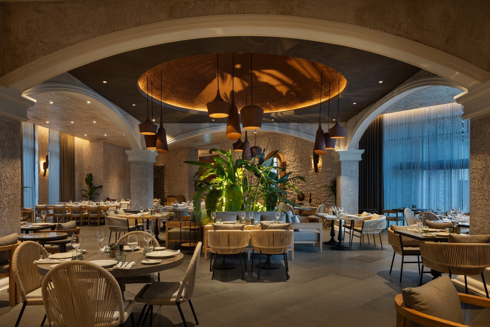 Restaurant des Hotels Fortuna of Cappadocia mit runden gedeckten Tischen und geflochtenen Stühlen unter Rundbögen. In der Mitte steht eine große Grünpflanze unter einem beleuchteten Deckenrund.