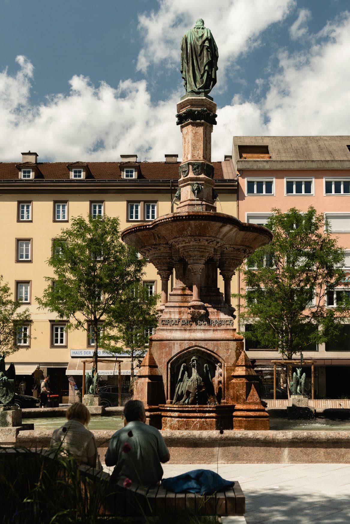 Der Rudolfsbrunnen am Bozner Platz in Innsbruck mit achteckiger Säule aus rotem Marmor und einer Bronzefigur auf der Spitze. Im Vordergrund sitzen zwei Personen mit dem Rücken zur Kamera am Beckenrand, dahinter Wohnhäuser und junge Bäume.