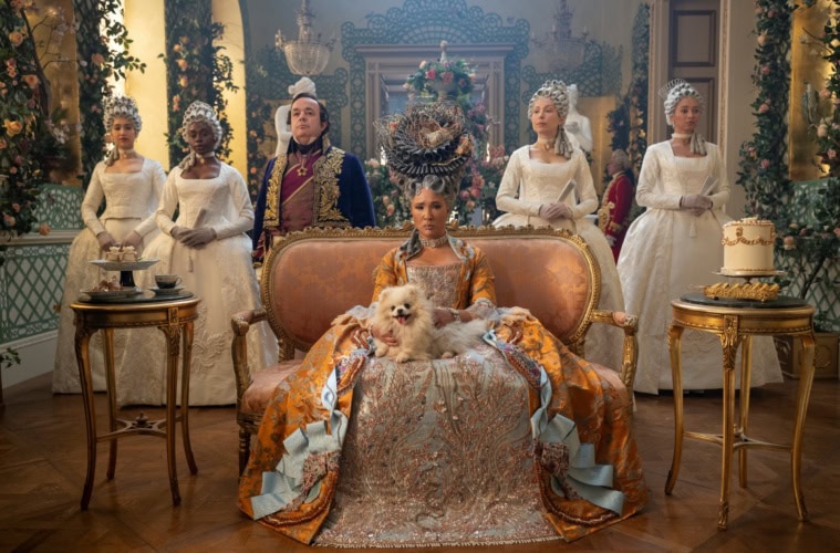 Still aus der Serie »Bridgerton«; Auf einem kleinen Sofa sitzt eine Frau in gold-silbernen Kleid mit einem weißen Hudn auf dem Schoß und blickt in die Kamera. Hinter ihr stehen aufgereiht vier Frauen in weißen Kleidern und ein Mann mit blau-goldenem Anzug.