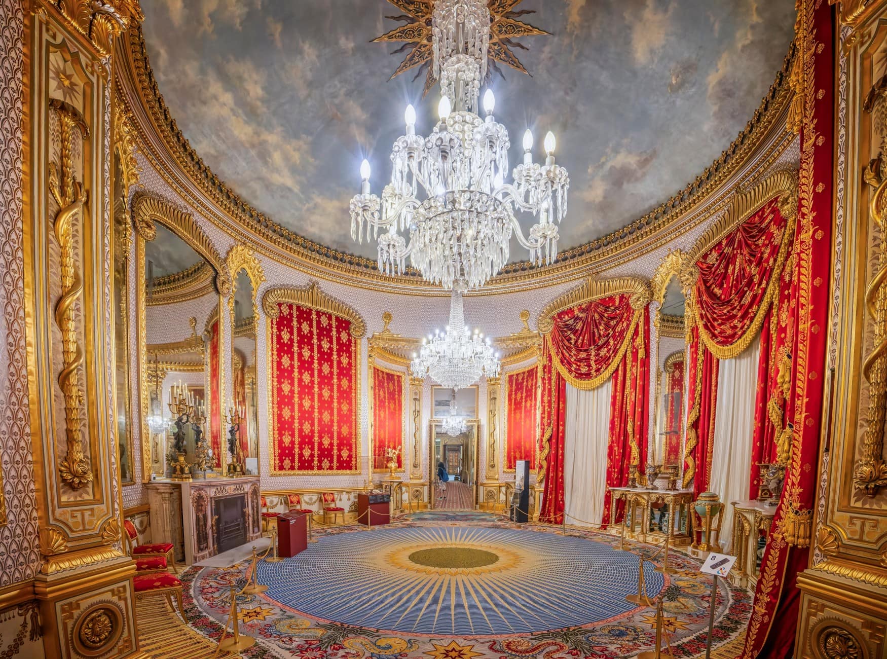 Ein kreisrunder Raum mit blauem Teppich, roten Wandbehängen zwischen goldenen Säulen und einem riesigen Kronleuchter in der Mitte.