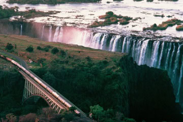 Luftaufnahme der Victoriafälle in Afrika; davor fährt ein Luxuszug von links nach rechts durchs Bild.