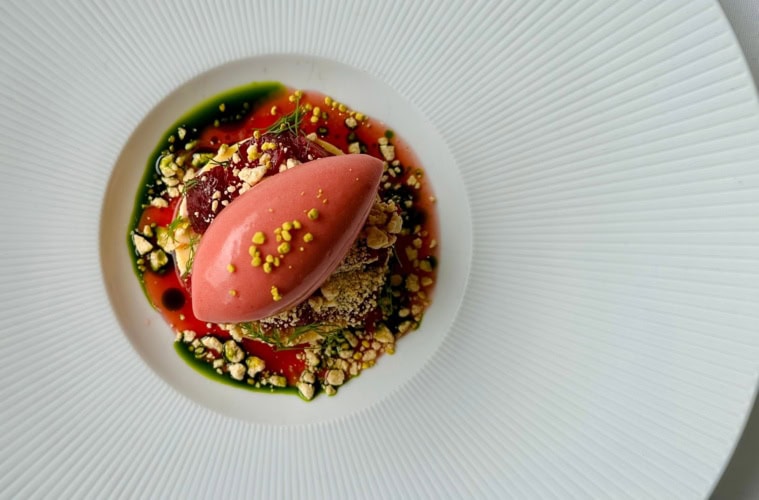 Auf einem schicken Teller mit sehr breitem Rand ist eine ovale, lachsfarbene Creme auf einem kleinen Bett aus Gebäck angerichtet, darunter Crumble und rote Sauce.