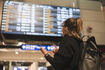Eine Frau steht vor einem Abflugsterminal in einem Flughafen und gleicht die Daten mit ihrem Smartphone ab.