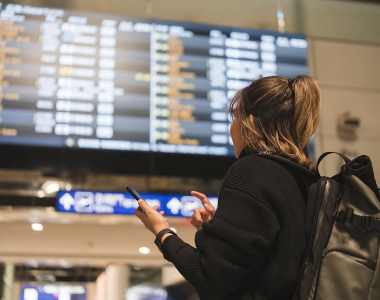 Eine Frau steht vor einem Abflugsterminal in einem Flughafen und gleicht die Daten mit ihrem Smartphone ab.