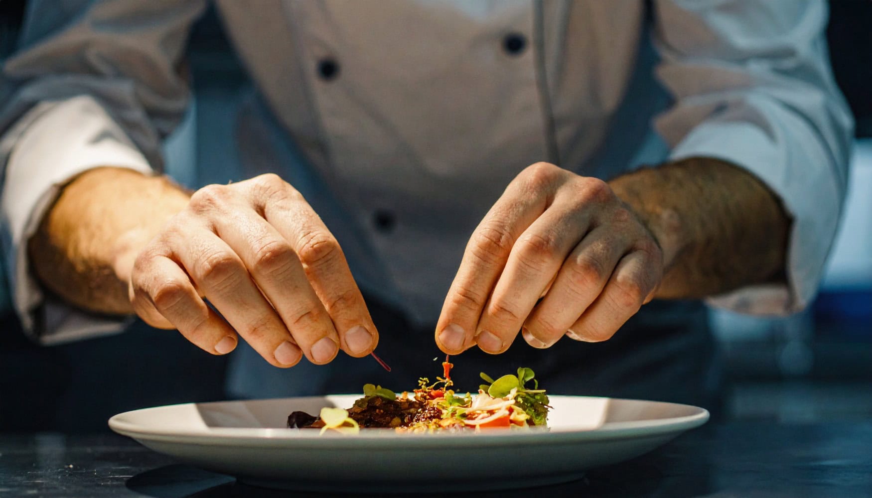 Nahaufnahme der Hände eines Kochs, der ein Gericht in einem Fine-Dining-Restaurant auf dem Teller anrichtet.