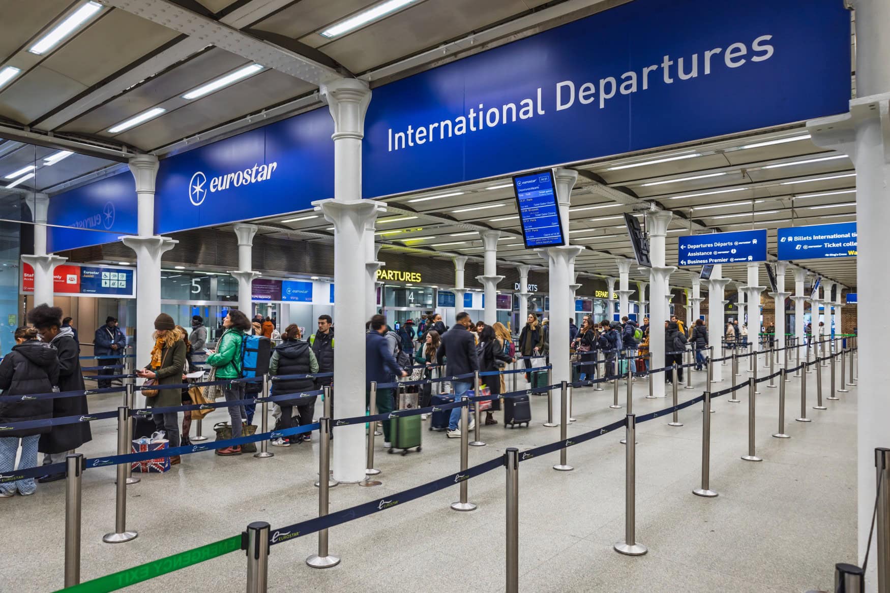 Eine geordnete Warteschlange mit Zuggästen und ihrem Gepäck, abgetrennt durch Bänder. Über den Menschen ist ein breites, blaues Schild mit der Aufschrift "eurostar" und "International Departures" angebracht.