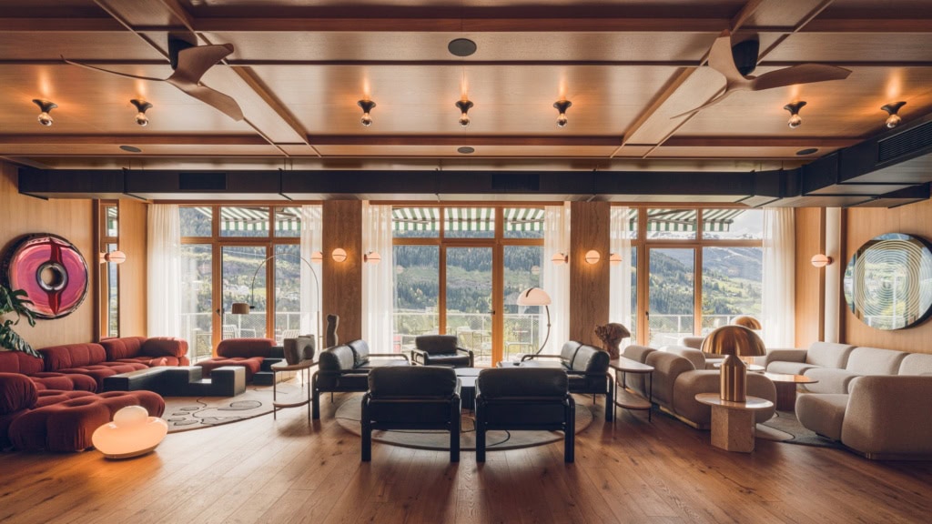 Eine weite, mit viel Holz verkleidete Hotellounge mit 50er-Jahre-Polstermöbeln. Das breite Fenster öffnet den Blick auf einen bewaldeten Berghang.