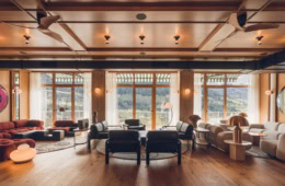 Eine weite, mit viel Holz verkleidete Hotellounge mit 50er-Jahre-Polstermöbeln. Das breite Fenster öffnet den Blick auf einen bewaldeten Berghang.