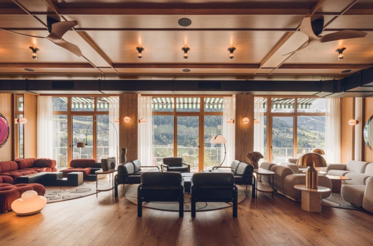 Eine weite, mit viel Holz verkleidete Hotellounge mit 50er-Jahre-Polstermöbeln. Das breite Fenster öffnet den Blick auf einen bewaldeten Berghang.