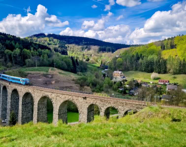 Ein blauer Zug fährt über ein Viadukt durch eine sommergrüne Landschaft.