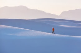 Ein einzelner Mann in roter Jacke, der inmitten der weiße Sanddünen vom Withe Sands Nationalpark spaziert, die im Licht fast blau wirken. Im Hintergrund sind die Umrisse von Felsen und Bergen zu erkennen, aber ganz blass und zart.