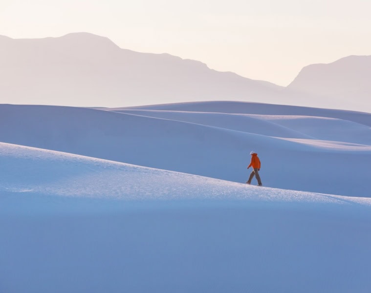 Ein einzelner Mann in roter Jacke, der inmitten der weiße Sanddünen vom Withe Sands Nationalpark spaziert, die im Licht fast blau wirken. Im Hintergrund sind die Umrisse von Felsen und Bergen zu erkennen, aber ganz blass und zart.