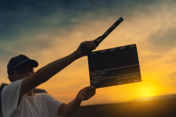 Junger Mann hält eine Filmklappe hoch. Im HIntergrund geht die Sonne unter.