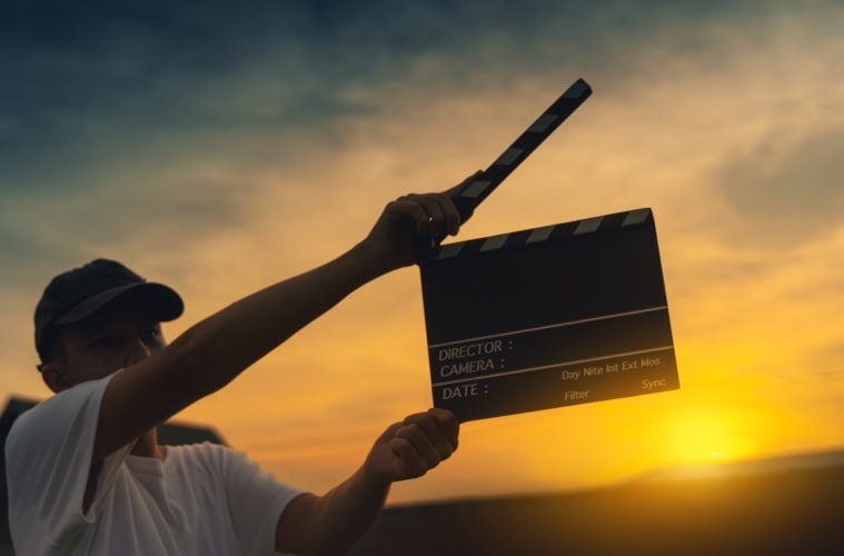 Junger Mann hält eine Filmklappe hoch. Im HIntergrund geht die Sonne unter.