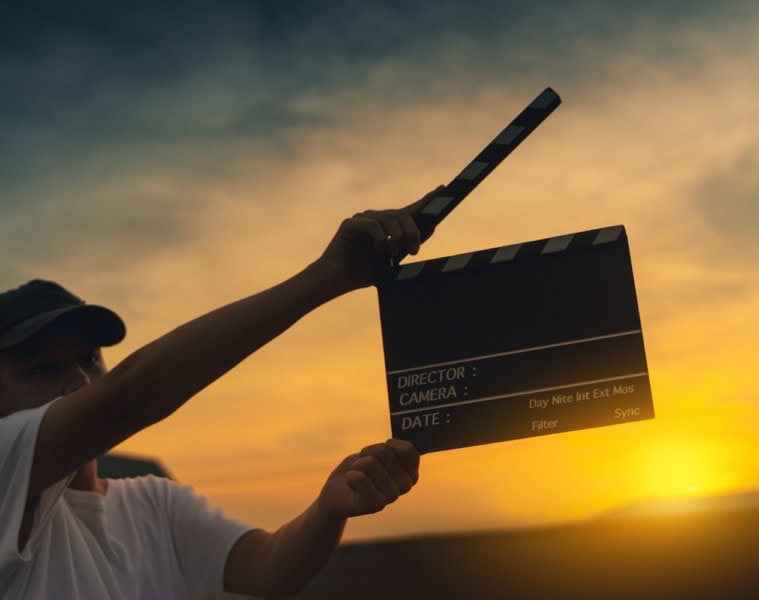 Junger Mann hält eine Filmklappe hoch. Im HIntergrund geht die Sonne unter.