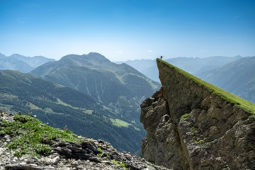 Panoramabild auf die Osttiroler Berge mit blauem Himmel: Auf einem markanten Berg, der spitz zur Seite ragt, steht auf dem Gipfel eine einzelne Person und reckt die Hände in den Himmel. Von der Fotoperspektive aus sieht sie sehr klein aus.