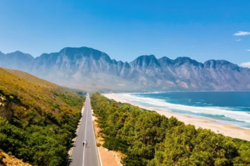 Luftaufnahme einer leeren Straße am Kogelbay Beach Südafrika, auf der zwei Fussgänger zu sehen sind. Im Hintergrund hohe Berge und blauer Himmel.