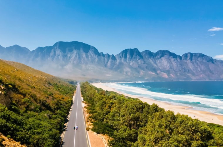 Luftaufnahme einer leeren Straße am Kogelbay Beach Südafrika, auf der zwei Fussgänger zu sehen sind. Im Hintergrund hohe Berge und blauer Himmel.