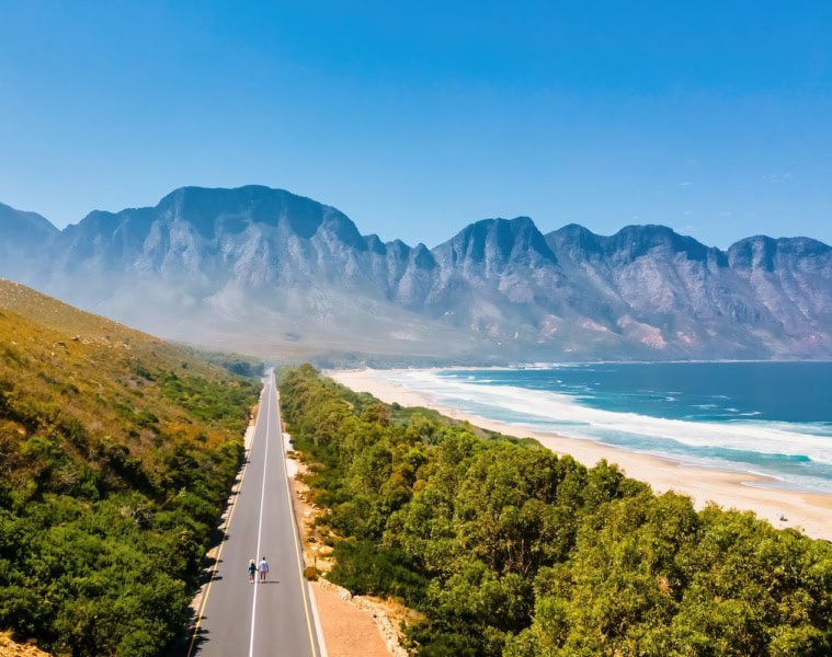 Luftaufnahme einer leeren Straße am Kogelbay Beach Südafrika, auf der zwei Fussgänger zu sehen sind. Im Hintergrund hohe Berge und blauer Himmel.