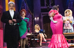 Musicalbühne mit verschiedenen Darstellern: Links ein grauhaariger Mann im Frack, der grimmig schaut, daneben eine singende Drag im lila-grünen Cocktailkleid, in der Mitte ein Bistrotisch mit Gläsern, an dem eine blonde Drag im rüschigen Kleid und langen übereinander geschlagenen Beinen sitzt. In der Mitte eine PoC-Dame im opulent pinken Abendkleid mit weißen langen Handschuhen, die sich mit einer weitere Drag, in ein biederes Kleid mit Schürze unterhält.
