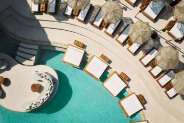 Luftaufnahme eines geschwungenen Pools in einem Resort auf Zypern. Am Rand ragen einige breite Liegen über die Wasseroberfläche.