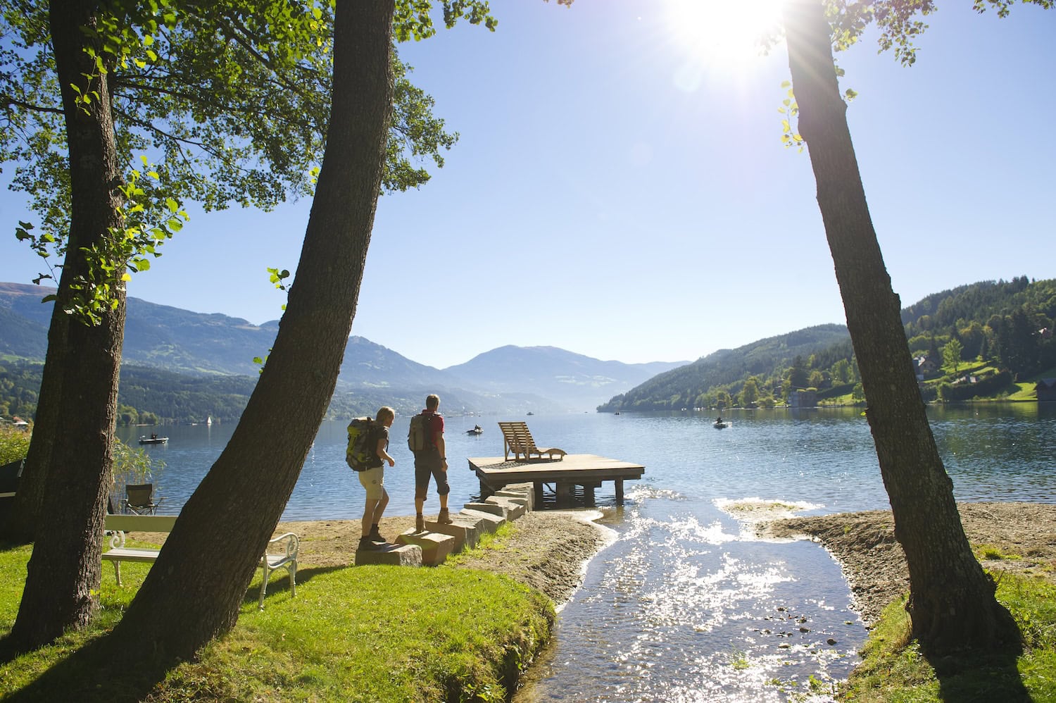 Zwei Wanderer blicken auf den Millstätter See vom Ufer aus. Im Vordergrund drei Bäume, die das Foto einrahmen. Vor den Wanderern ist ein kleiner Steg mit einem Holzliegestuhl, auf dem See sind drei Botte und im Hintergrund die Berge. Die Sonne scheint und es ist ein schöner Sommertag im Urlaub.
