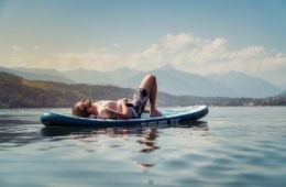 Mann liegt auf seinem Stand-Up-Paddle in der Sonne und entspannt auf dem Millstätter See im Sommer. Im Hintergrund Berge und weisse Wolken.