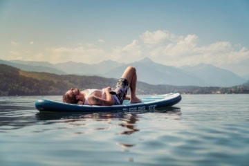 Mann liegt auf seinem Stand-Up-Paddle in der Sonne und entspannt auf dem Millstätter See im Sommer. Im Hintergrund Berge und weisse Wolken.