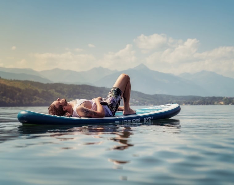 Mann liegt auf seinem Stand-Up-Paddle in der Sonne und entspannt auf dem Millstätter See im Sommer. Im Hintergrund Berge und weisse Wolken.