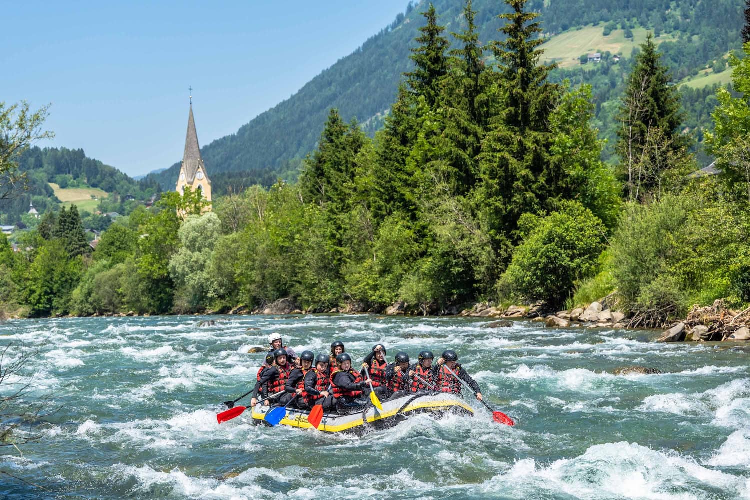 Wildwasser-Rafting, ein Boot mit 11 Menschen, die auf einem sprudelnden Fluss paddeln. Im Hintergrund lugt zwischen dichten Bäumen ein Kirchturm auf, darum herum Berge mit dichten Bäumen und einer Albwiese.