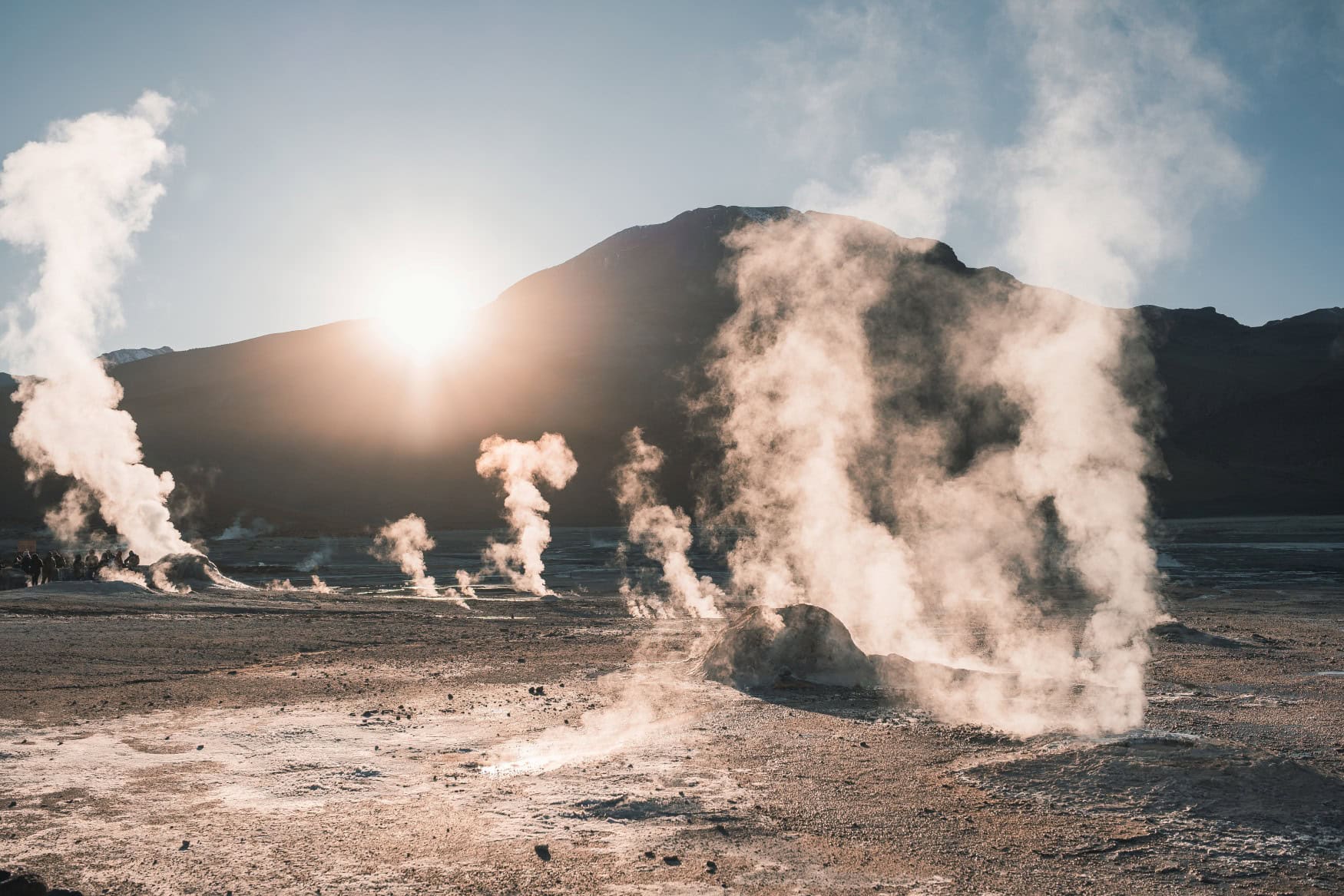 In der Atacama-Wüste steigt aus zahlreichen Stellen im Boden weißer Rauch auf. Im Hintergrund geht die Sonne hinter einem Berg unter.