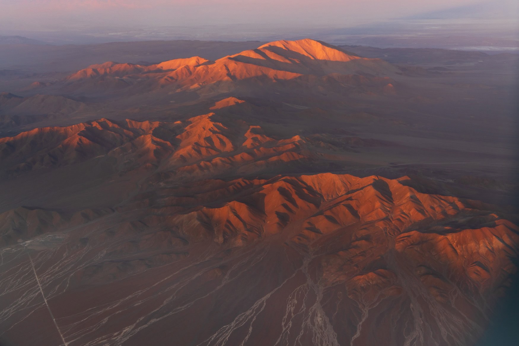 Luftbild der Atacama-Wüste in Chile; nur die Bergkuppen der Anden werden von der Morgensonne rot angestrahlt, der Rest der kargen Landschaft liegt im Schatten.