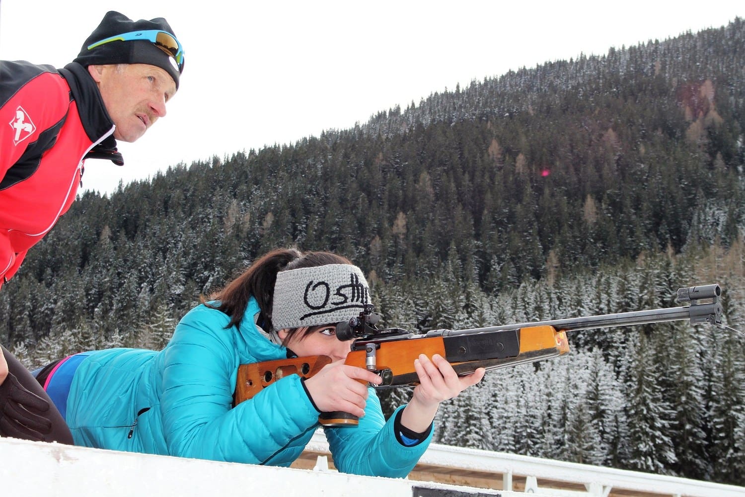 Frau liegt beim Biathlon-Schnuppertraining in Obertilliach in Osttirol mit dem Gewehr am Schießstand, daneben ein Trainer