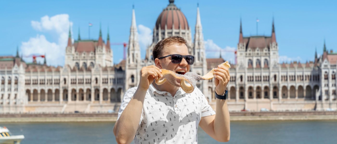 Ein Mann in sommerlicher Kleidung isst genüsslich ein Langos, hinter ihm die Donau und das Parlament in Budapest.