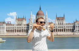 Ein Mann in sommerlicher Kleidung isst genüsslich ein Langos, hinter ihm die Donau und das Parlament in Budapest.