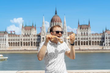 Ein Mann in sommerlicher Kleidung isst genüsslich ein Langos, hinter ihm die Donau und das Parlament in Budapest.