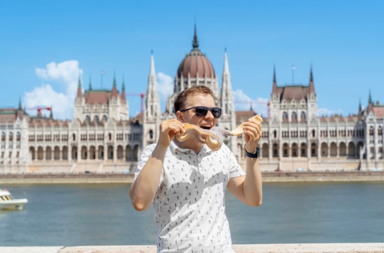 Ein Mann in sommerlicher Kleidung isst genüsslich ein Langos, hinter ihm die Donau und das Parlament in Budapest.