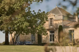 Sandsteinfassade des Château Cordeillan-Bages mit Eckturm und davor parkendem blauen Oldtimer-Cabrio im Schatten alter Bäume.