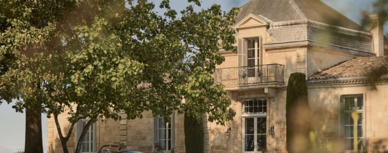 Sandsteinfassade des Château Cordeillan-Bages mit Eckturm und davor parkendem blauen Oldtimer-Cabrio im Schatten alter Bäume.