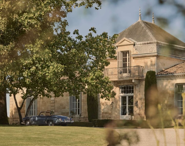 Sandsteinfassade des Château Cordeillan-Bages mit Eckturm und davor parkendem blauen Oldtimer-Cabrio im Schatten alter Bäume.