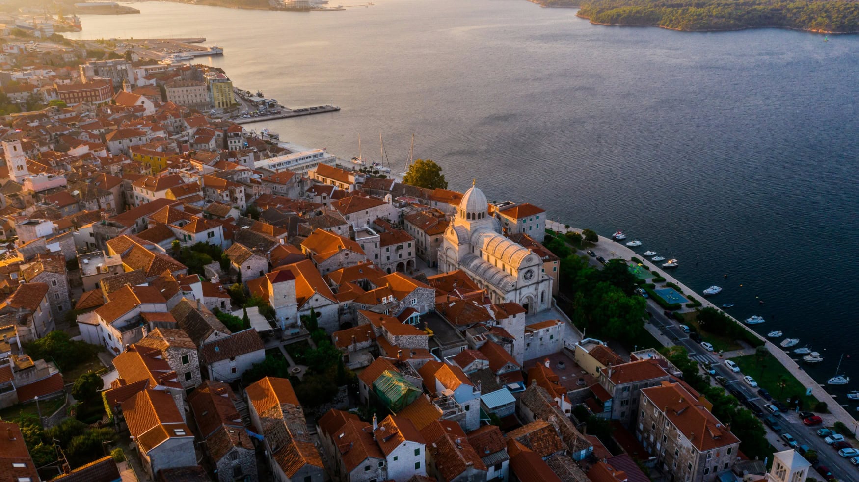 Luftansicht der mittelalterlichen Stadt Sibenik im kroatischen Dalmatien; rechts die Adria, mittig die Stadt mit weißer Kathedrale zwischen zahlreichen roten Dächern.