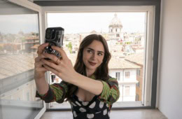 Junge Frau mit langen braunen Haaren macht ein Selfie mit Smartphone und kompakter Filmkamera am offenen Fenster, im Hintergrund die Dächer und eine Kirchenkuppel von Rom