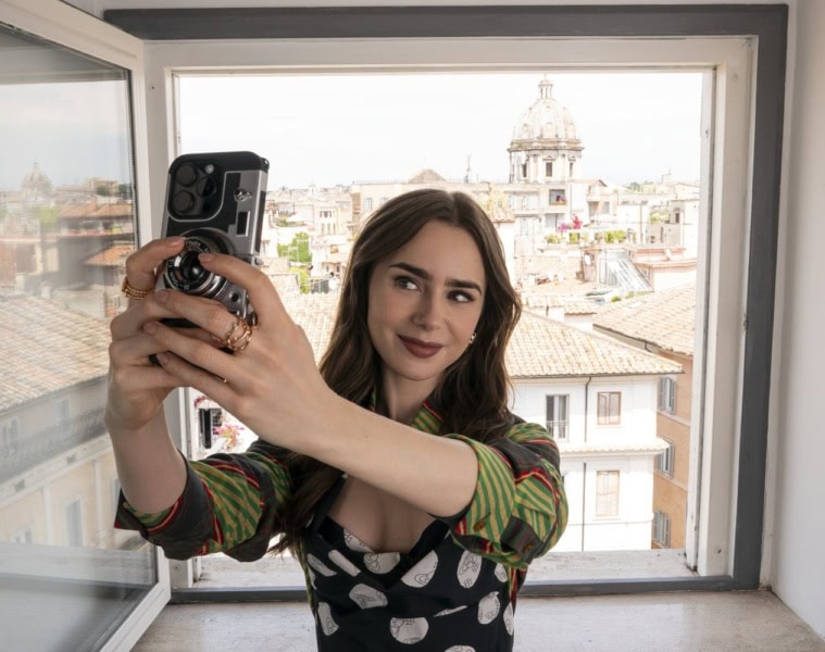 Junge Frau mit langen braunen Haaren macht ein Selfie mit Smartphone und kompakter Filmkamera am offenen Fenster, im Hintergrund die Dächer und eine Kirchenkuppel von Rom