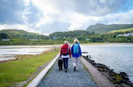 Ein von Stein umfasster grüner Pfad führt geradwegs über eine Meerenge auf eine Insel. Darauf wandern zwei Menschen in Outdoorkleidung. Im Hintergrund sind grüne Berge erkennbar.