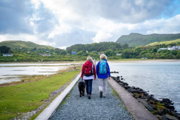 Ein von Stein umfasster grüner Pfad führt geradwegs über eine Meerenge auf eine Insel. Darauf wandern zwei Menschen in Outdoorkleidung. Im Hintergrund sind grüne Berge erkennbar.