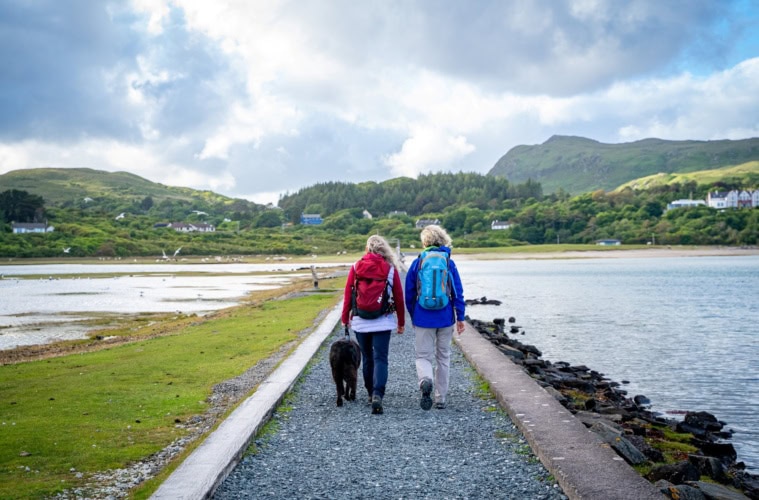 Ein von Stein umfasster grüner Pfad führt geradwegs über eine Meerenge auf eine Insel. Darauf wandern zwei Menschen in Outdoorkleidung. Im Hintergrund sind grüne Berge erkennbar.