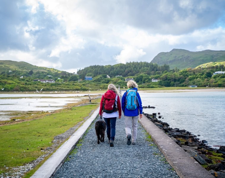 Ein von Stein umfasster grüner Pfad führt geradwegs über eine Meerenge auf eine Insel. Darauf wandern zwei Menschen in Outdoorkleidung. Im Hintergrund sind grüne Berge erkennbar.