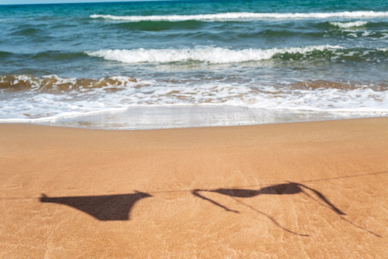 Auf dem Sand eines sommerlichen Strands ist der Schatten eines Bikinis an einer Wäscheleine zu sehen.